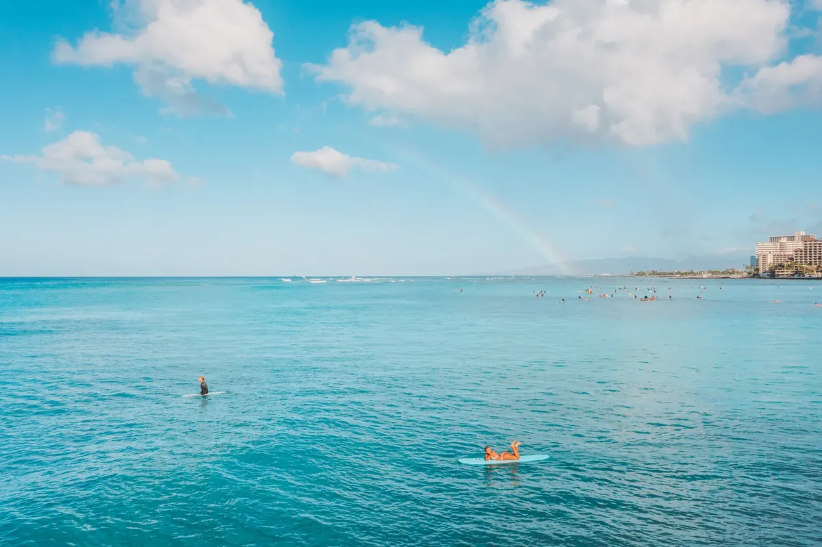 Surfers enjoy calm blue waters and a stunning rainbow over the horizon. Perfect weather for leisure and relaxation.